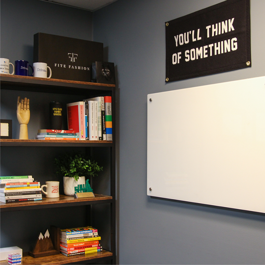 A modern office space featuring a dark wooden bookshelf filled with books, mugs, decorative items, and a box labeled "Fite Fashion." A whiteboard is mounted on the adjacent dark blue wall, with a black fabric banner above it that reads, "YOU'LL THINK OF SOMETHING." The setting has a creative and organized ambiance, with design and branding materials prominently displayed.